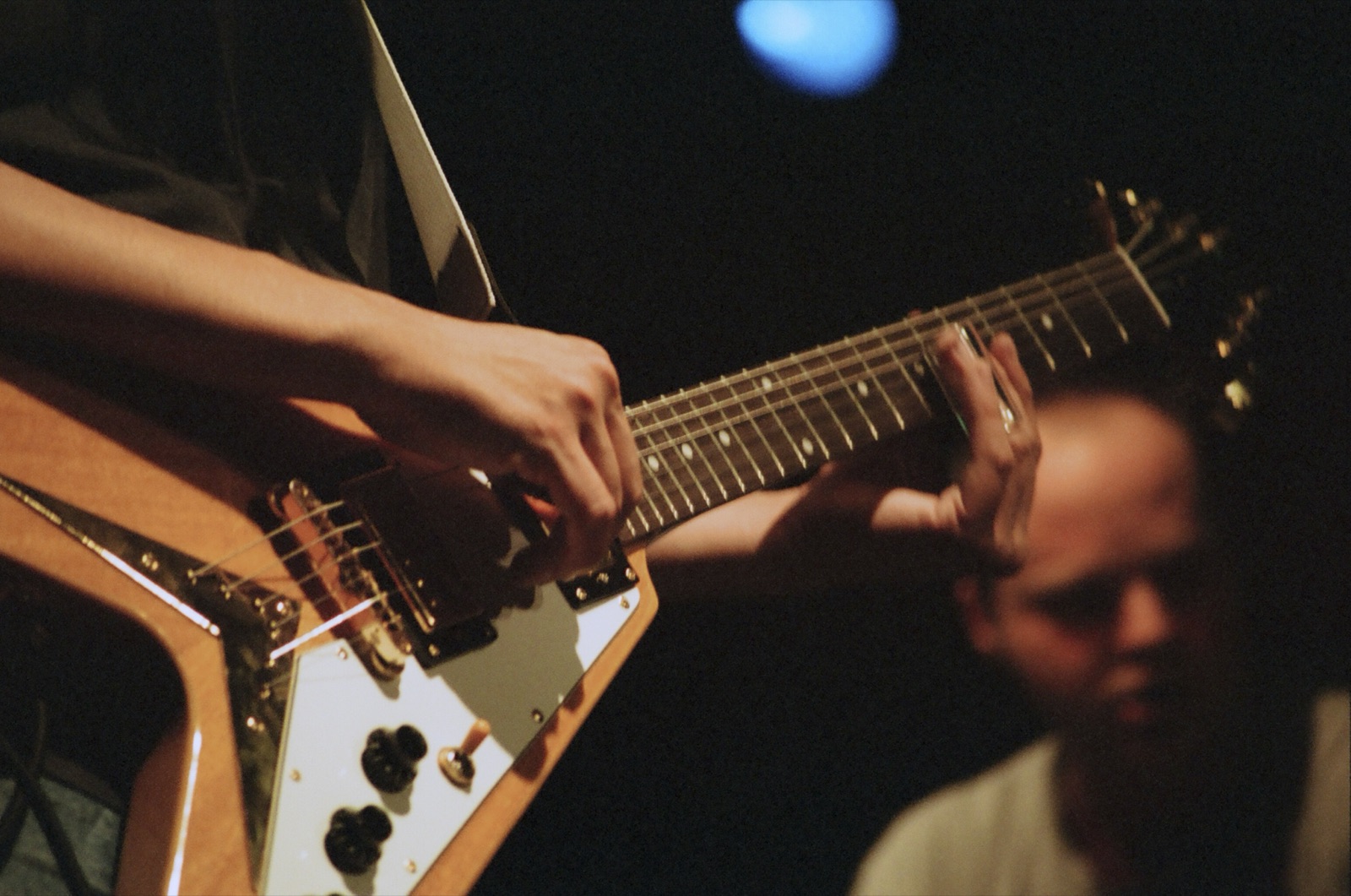 Close view of hands on a Flying V guitar neck, with the organist soft in the background and a blue stage light above.